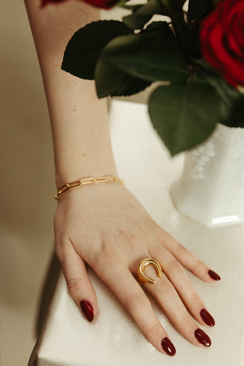 Hand with gold horseshoe ring and bracelet, red nail polish, and a vase of red roses.